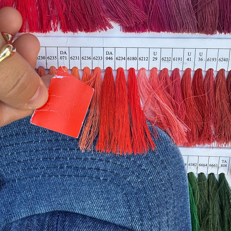 Color swatch being held up against different warm colored thread, and a denim cap.
