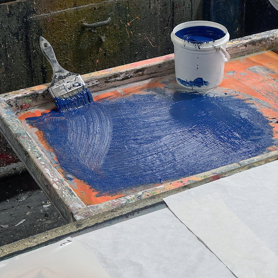 Paintbrush and bucket of blue paint on a wooden screenprint panel. 