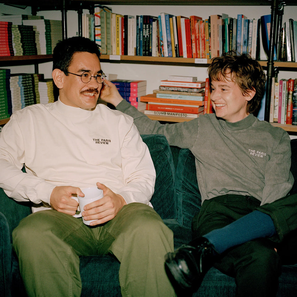 Two people sitting on a couch with bookshelves filled with books in the background wearing Long Sleeve Trash Tees with "The Paris Review" embroidered on the left chest area.