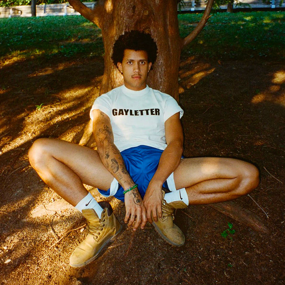 Person wearing a custom Tailored Trash Tee with 'GAYLETTER' text, sitting under a tree.