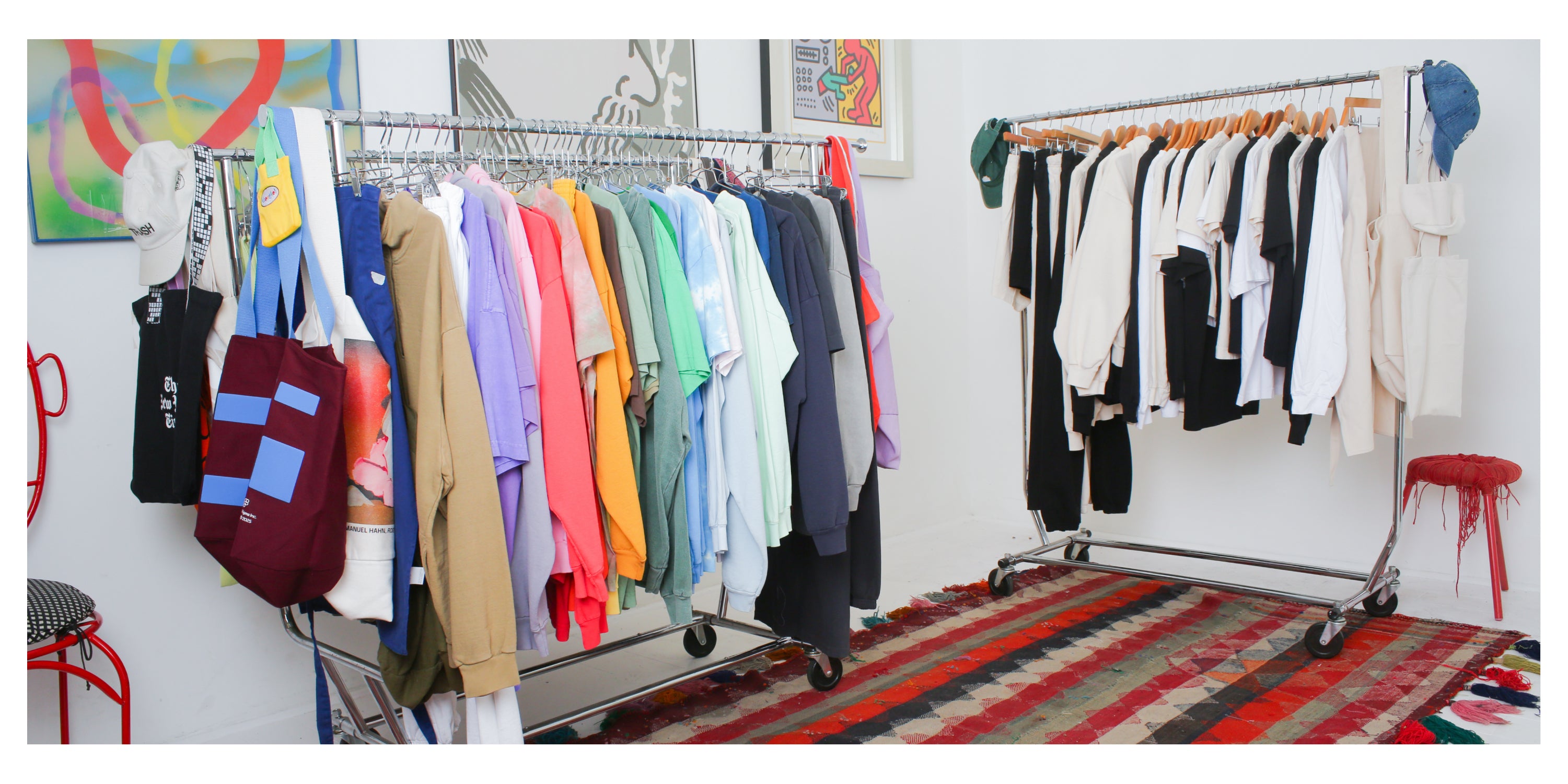 Two clothing racks, one with colorful custom clothing pieces, the other with Everybody.World garments in White, Black, and Scour. Colorful art and rug.