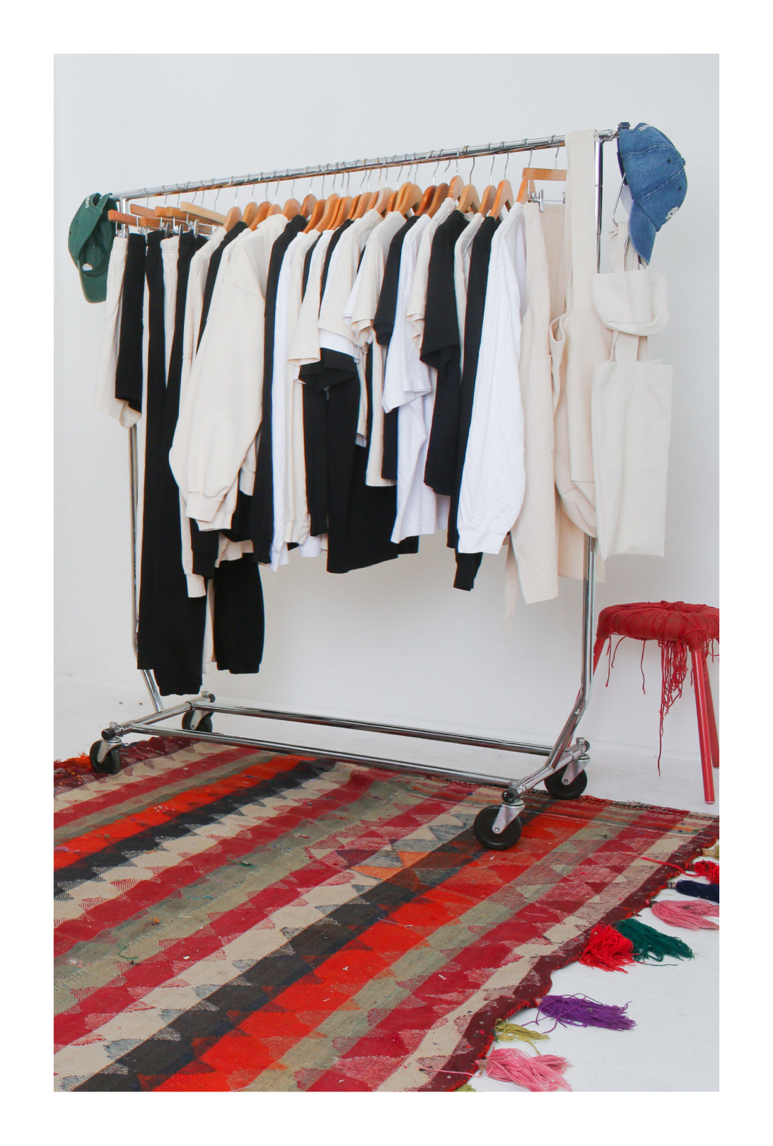 Clothing rack with Everybody.World garments in White, Black, and Scour. Colorful rug on the floor below. 