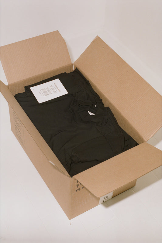 Large brown shipping box, open to show black tees folded inside with a notecard from Everybody.World.