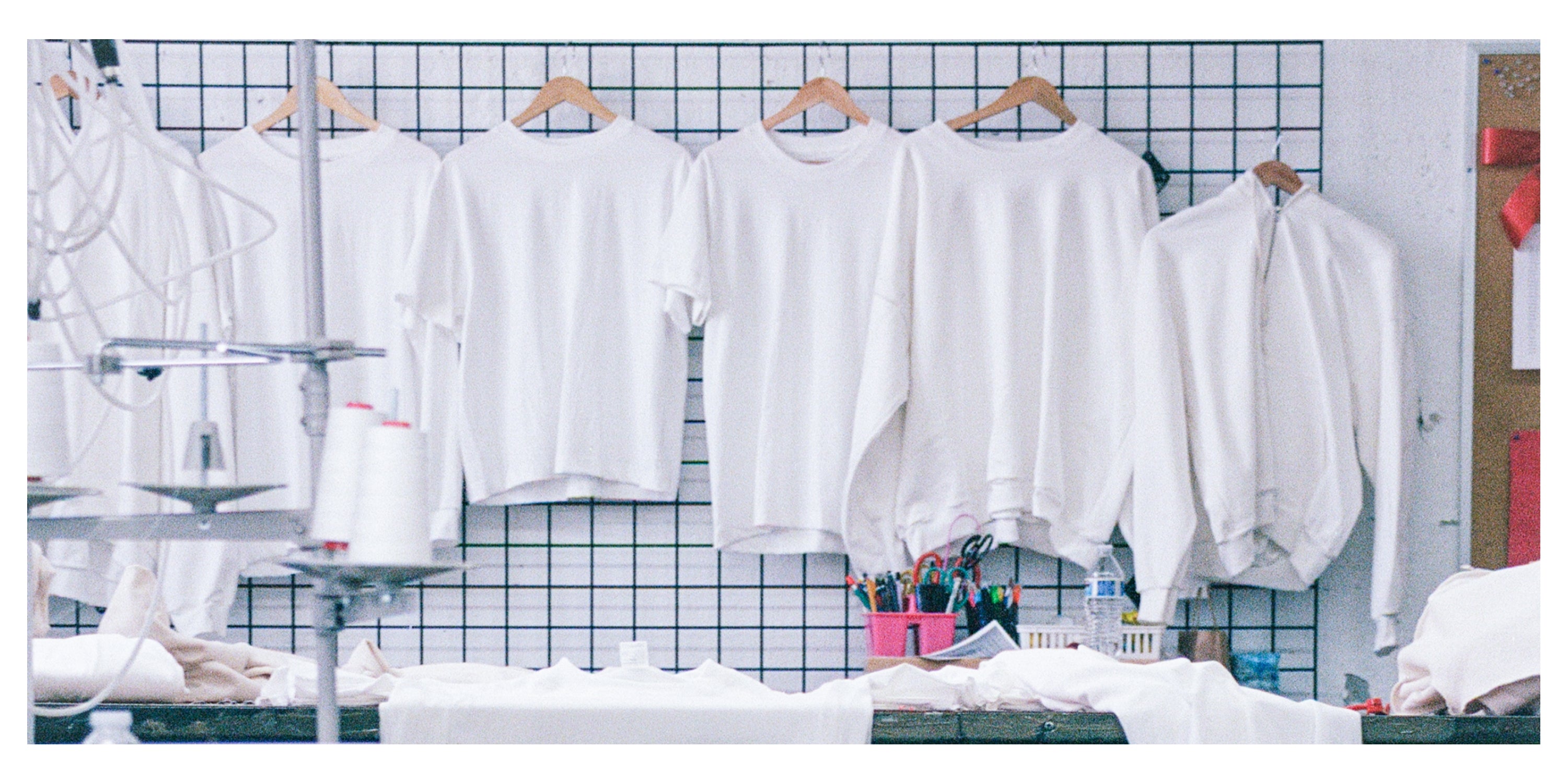 Everybody.World T-shirts and sweatshirts in pfd hanging on a wall grid with sewing machine parts in the foreground.
