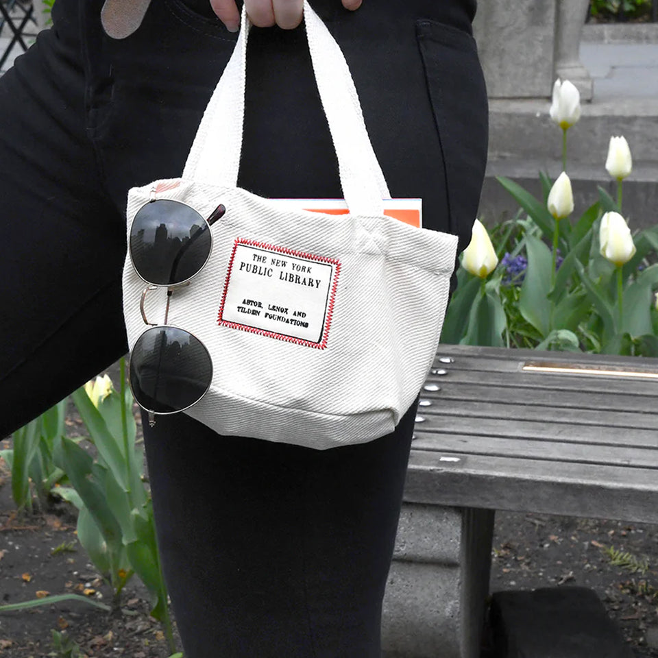 Person holding a custom "The New York Public Library" Mini Tote with sunglasses and a book, standing in a garden and bench.